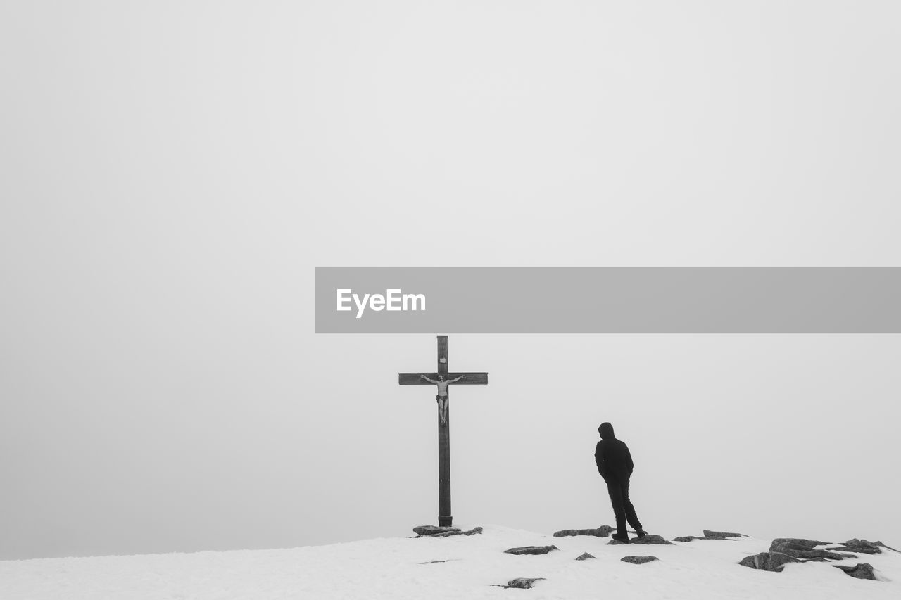 Man standing on snow covered hill by religious cross against sky during winter