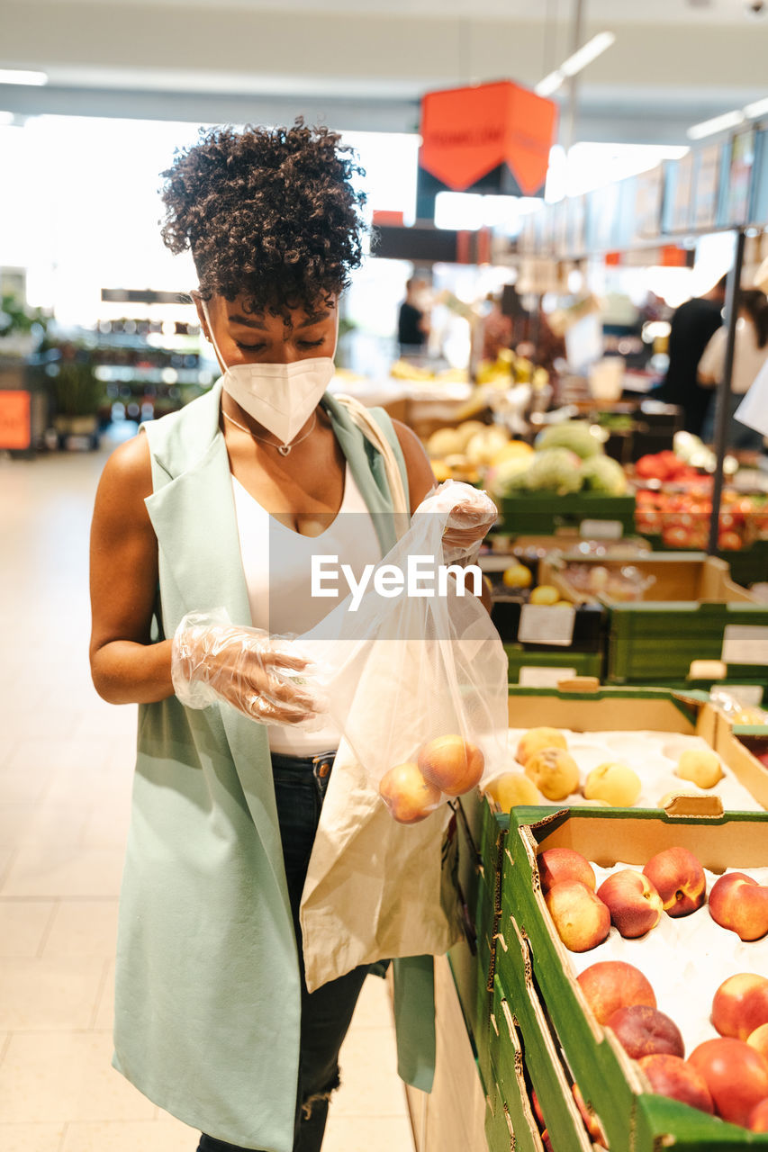 Side view of african american female customer in protective mask and gloves picking fruit from box while making purchases in supermarket during coronavirus pandemic