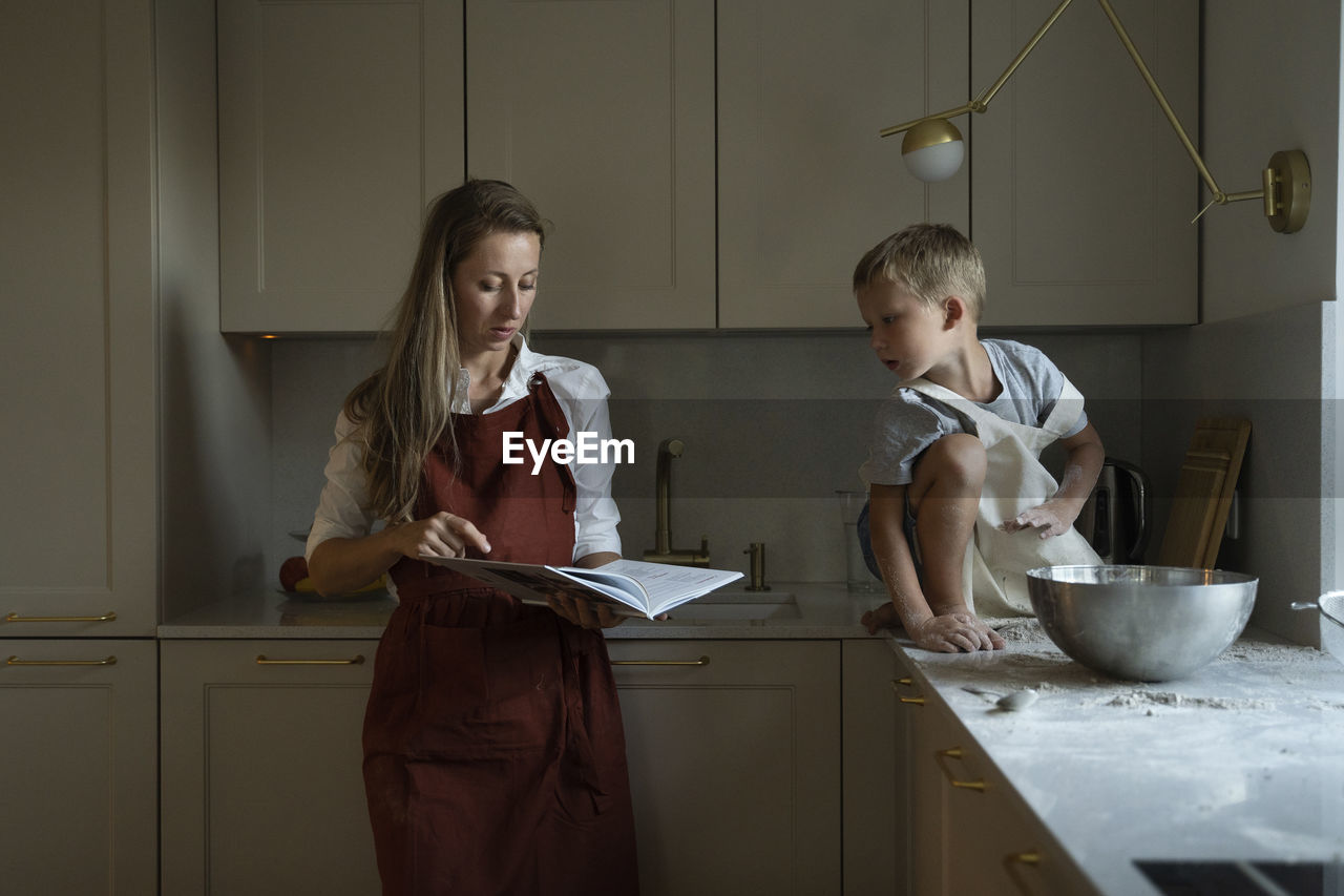 Mother reading recipe book with son sitting on kitchen counter