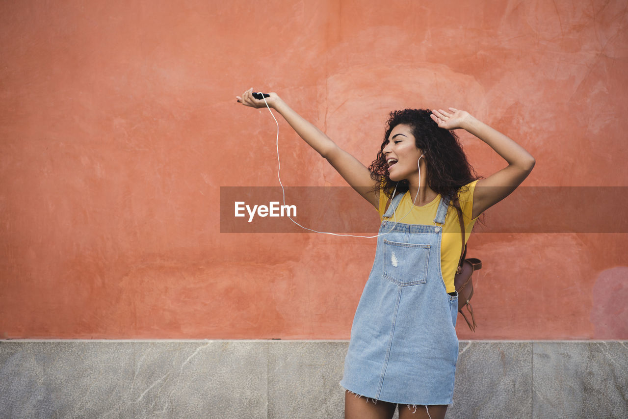 Cheerful woman listening to music on mobile phone while standing against wall