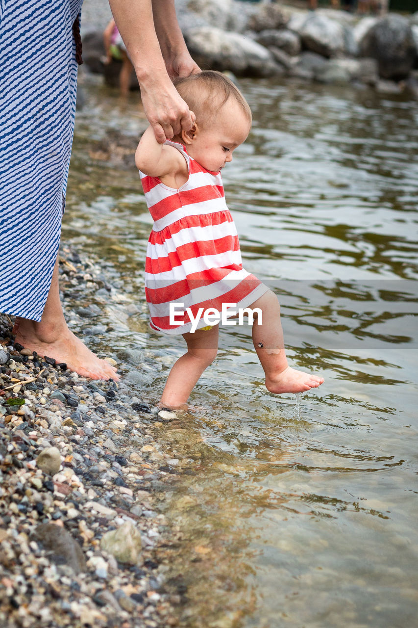 Close up view on baby in striped dress making steps in water being held by motherhands