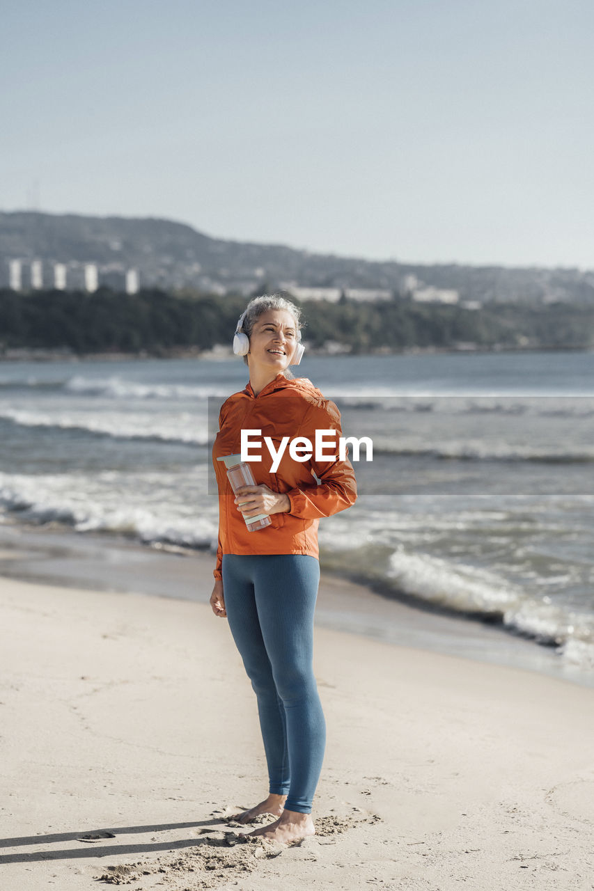 Smiling mature woman with water bottle standing on shore at beach