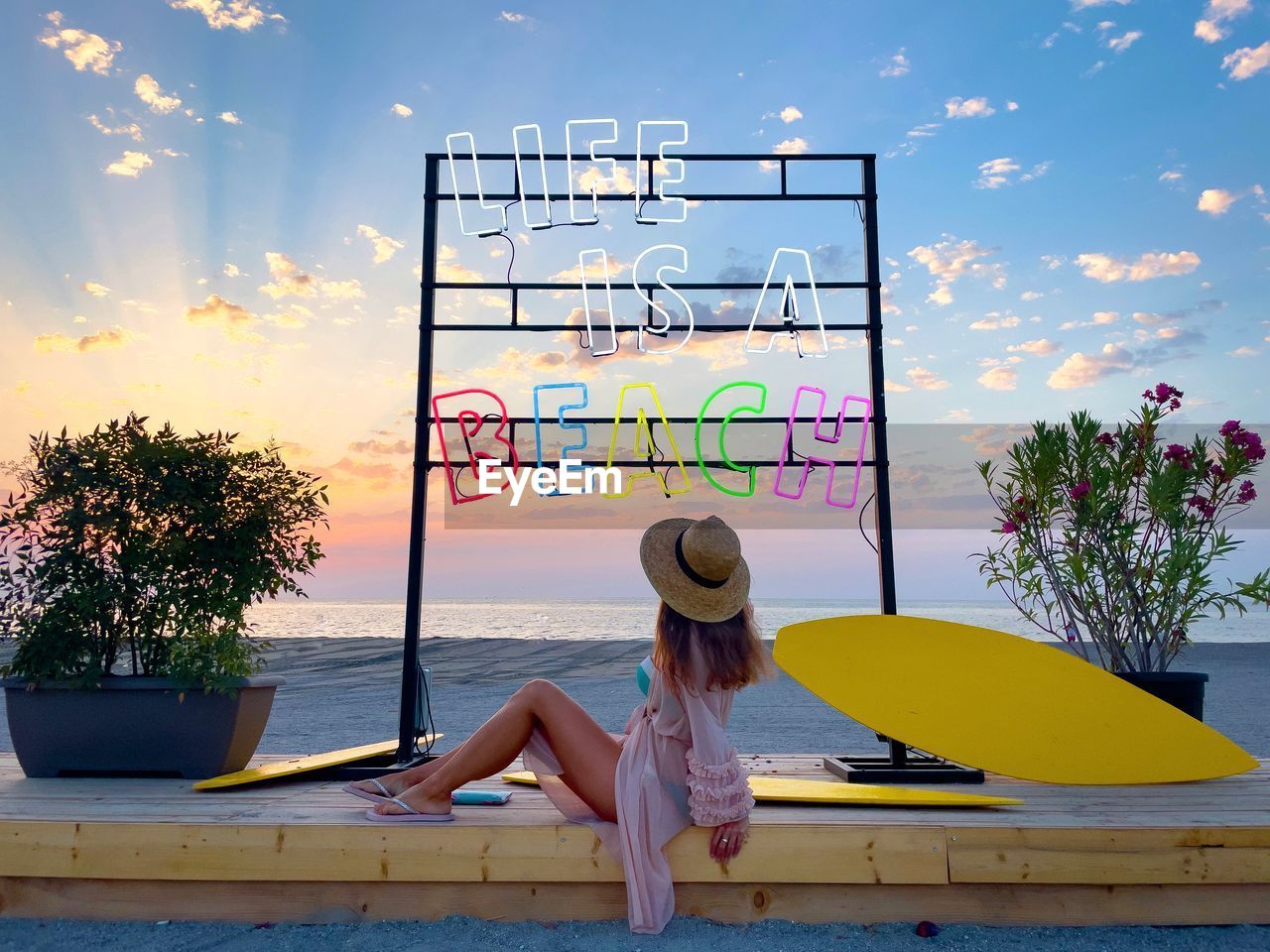 Fashionable young woman on the beach sitting under a colorful neon sign