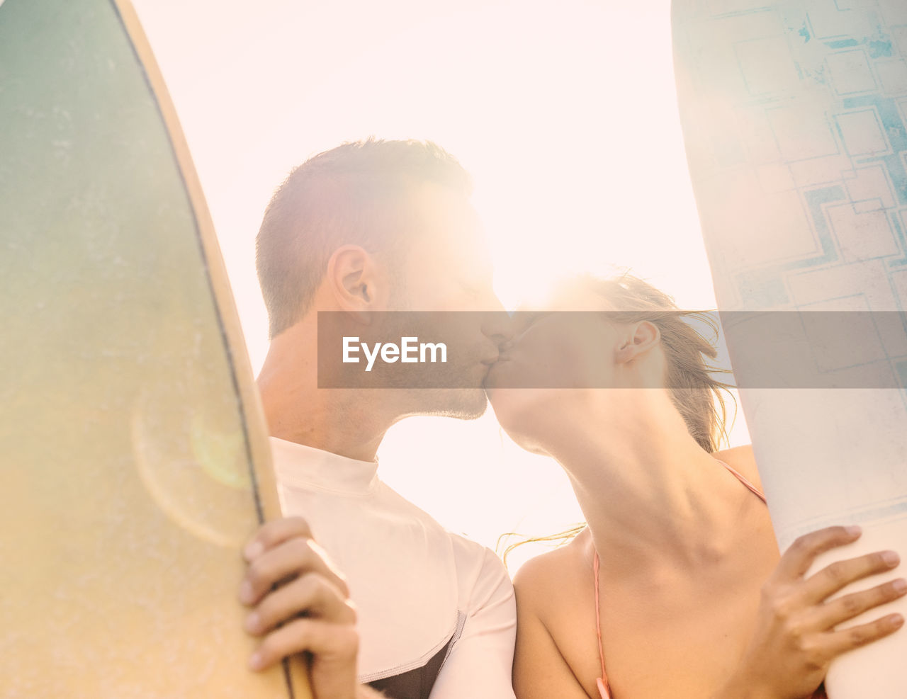 Low angle view of couple with surfboards kissing against sky during sunny day