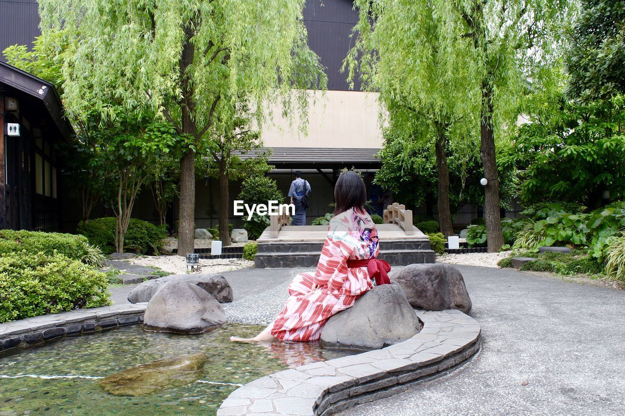 Rear view of woman relaxing on rock by pond against shrine