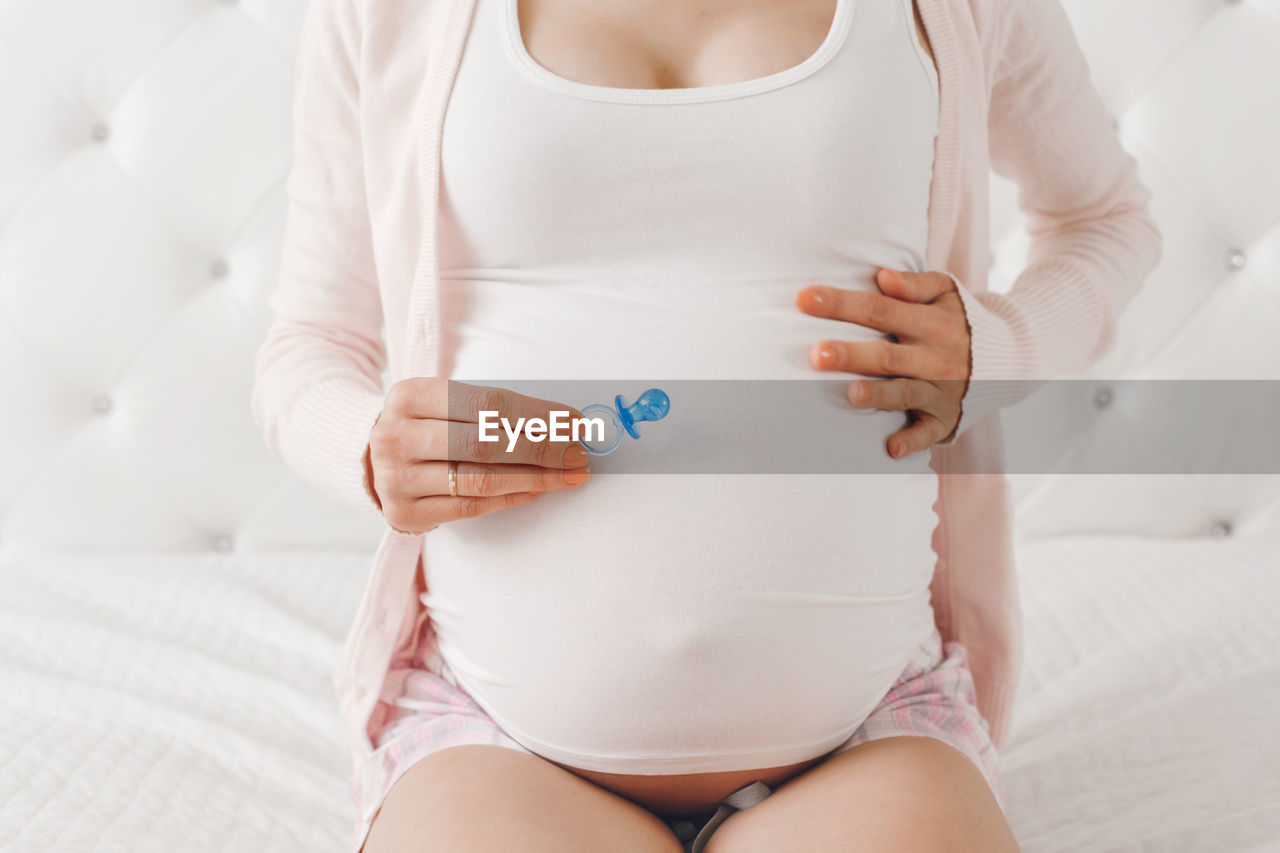 Midsection of pregnant woman holding pacifier sitting on bed at home