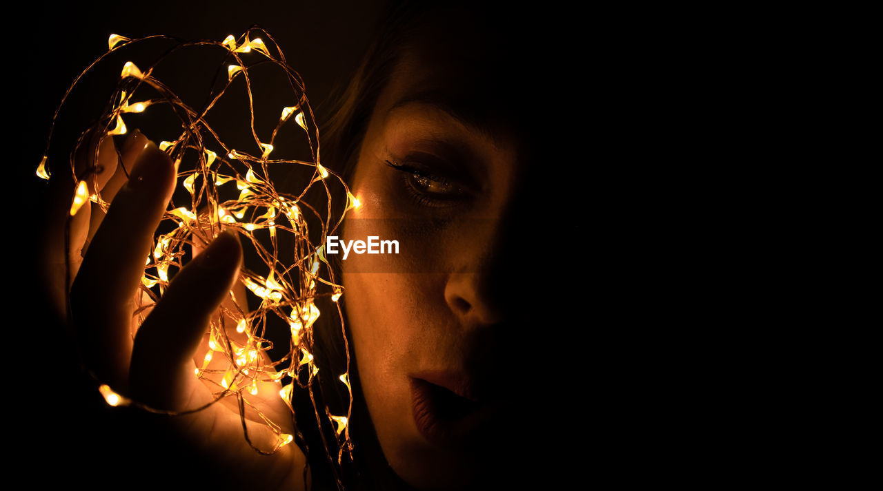 Close-up of woman holding christmas lights against black background