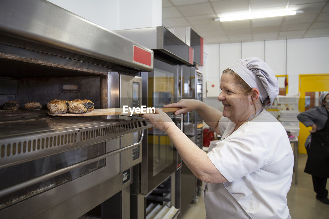 Side view of glad female baker in uniform taking out freshly baked bread from electric modern oven in baking school