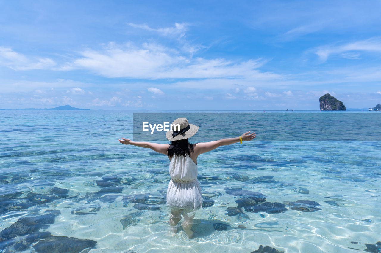 REAR VIEW OF WOMAN STANDING AT SEA SHORE AGAINST SKY