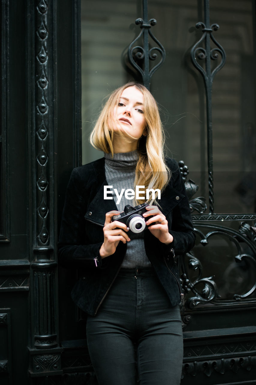 PORTRAIT OF YOUNG WOMAN HOLDING CAMERA WHILE STANDING IN KITCHEN