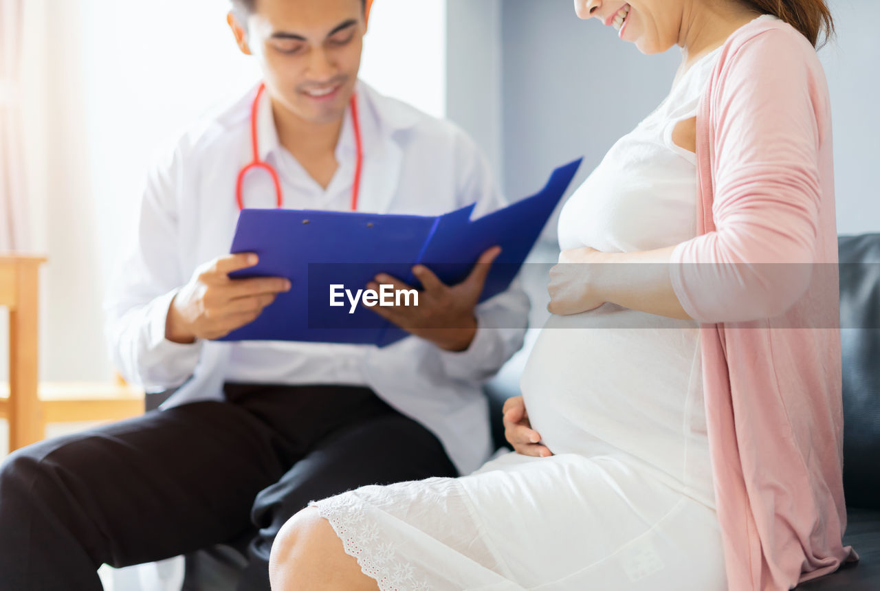 Doctor holding medical record while pregnant woman sitting at home