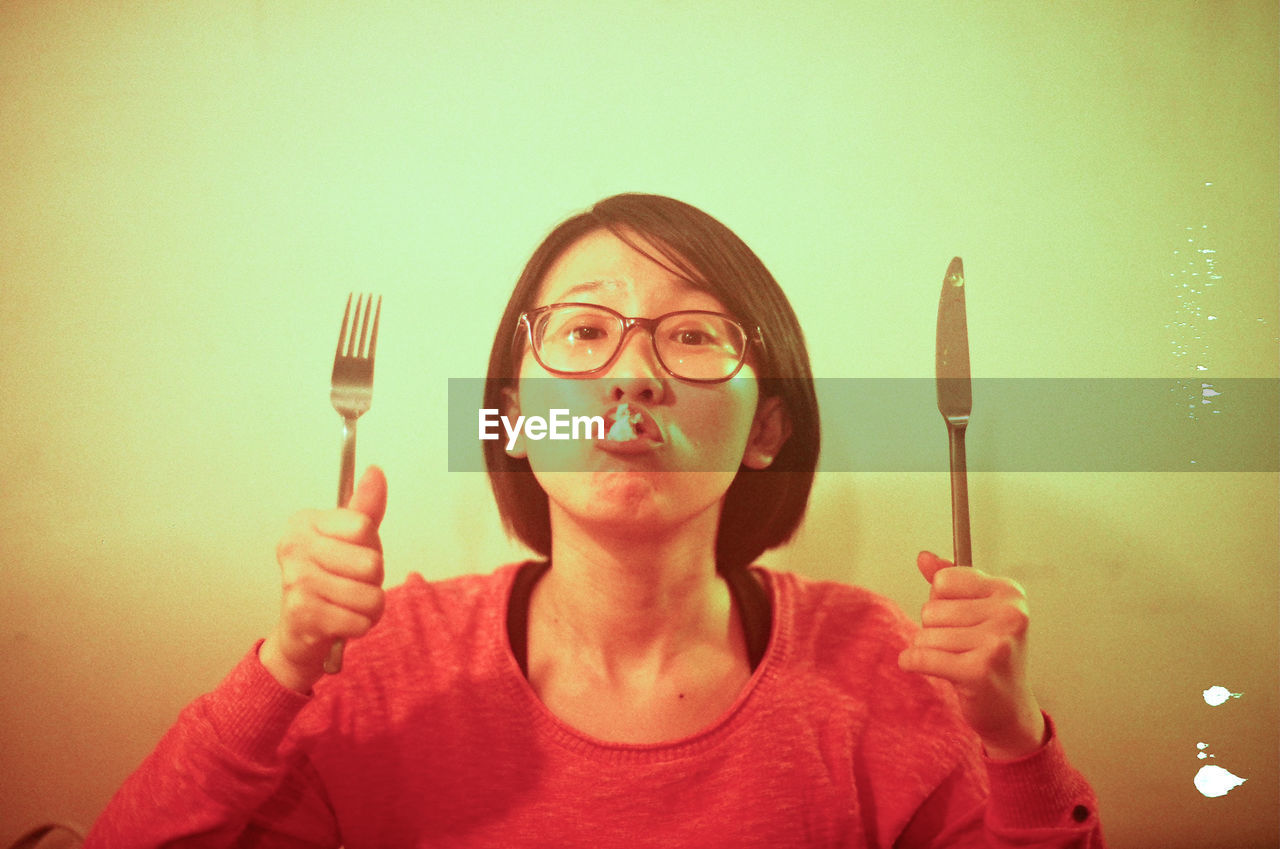 Portrait of young woman making face while holding fork and butter knife at home