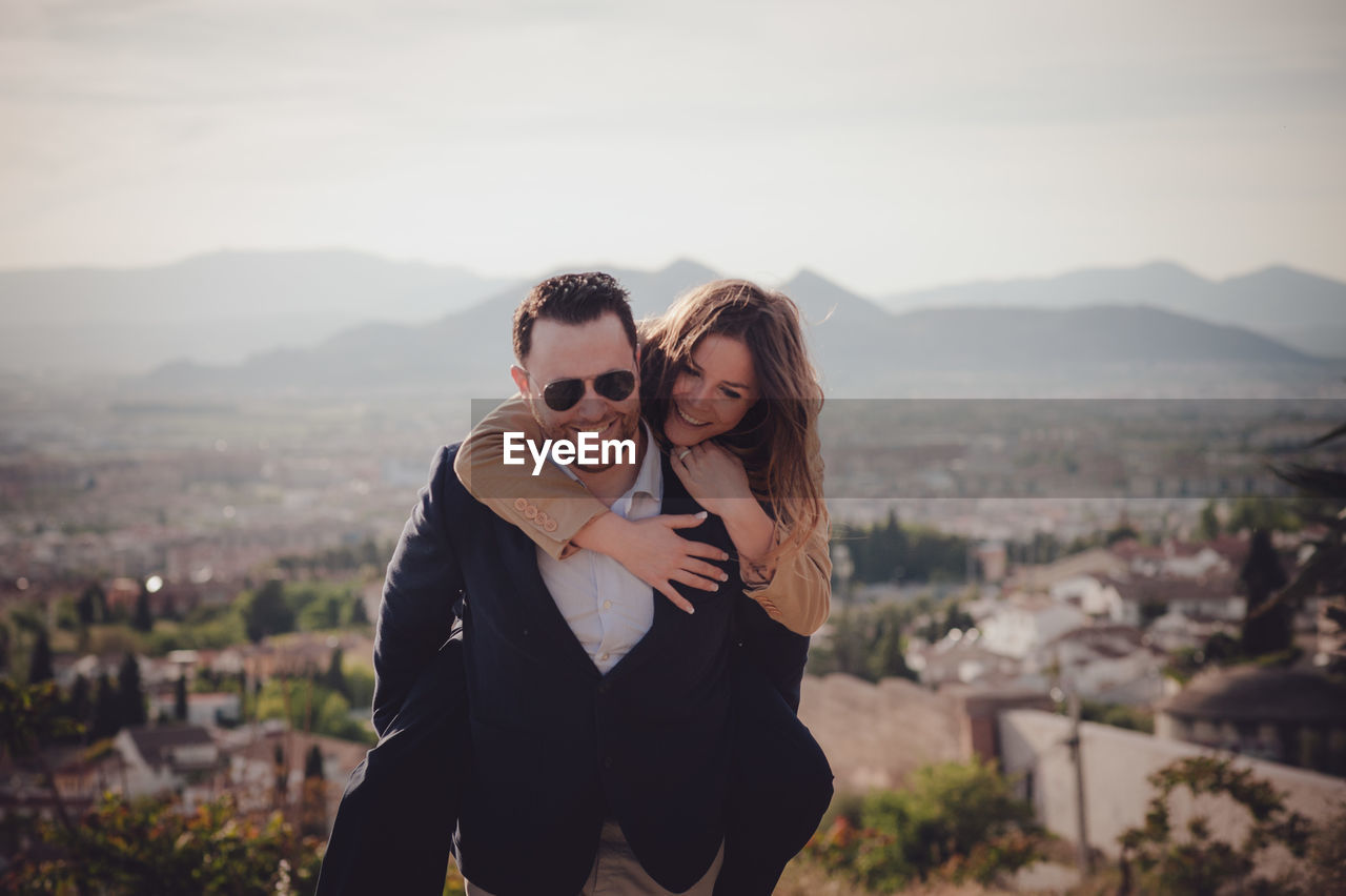 Man giving piggy back ride to happy woman standing against cityscape