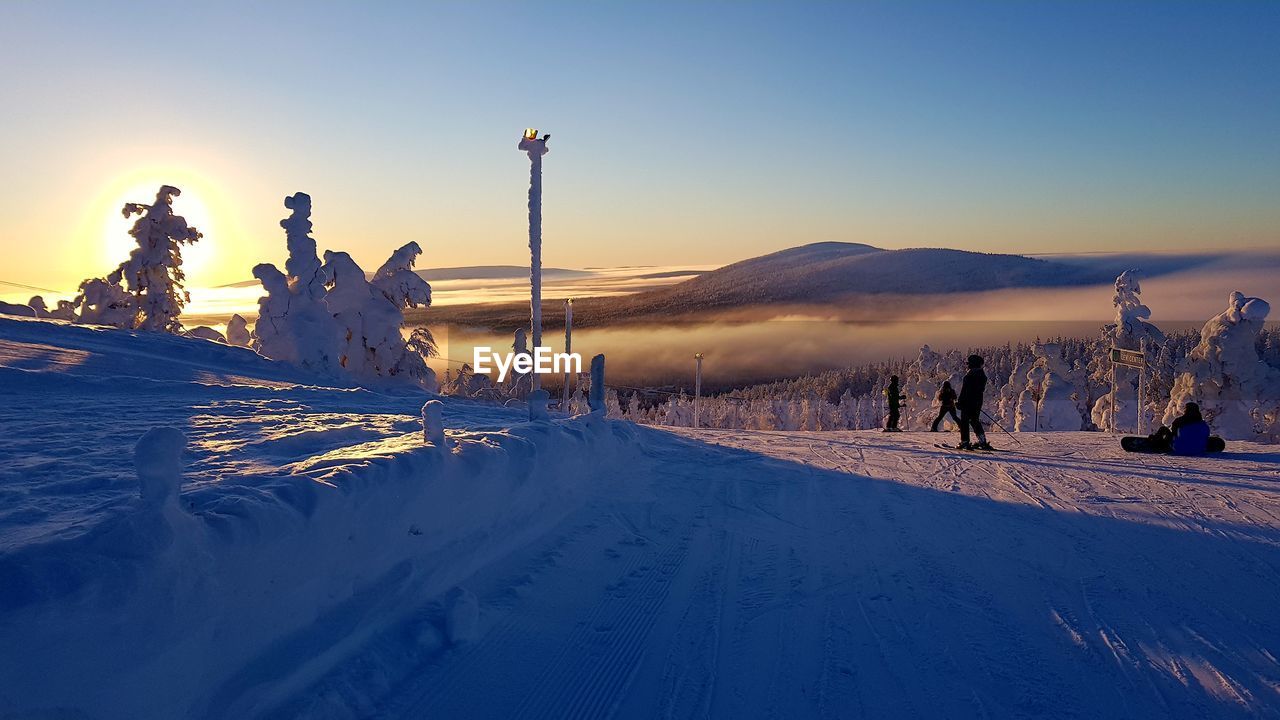 People skiing on snow covered field against sky during sunset