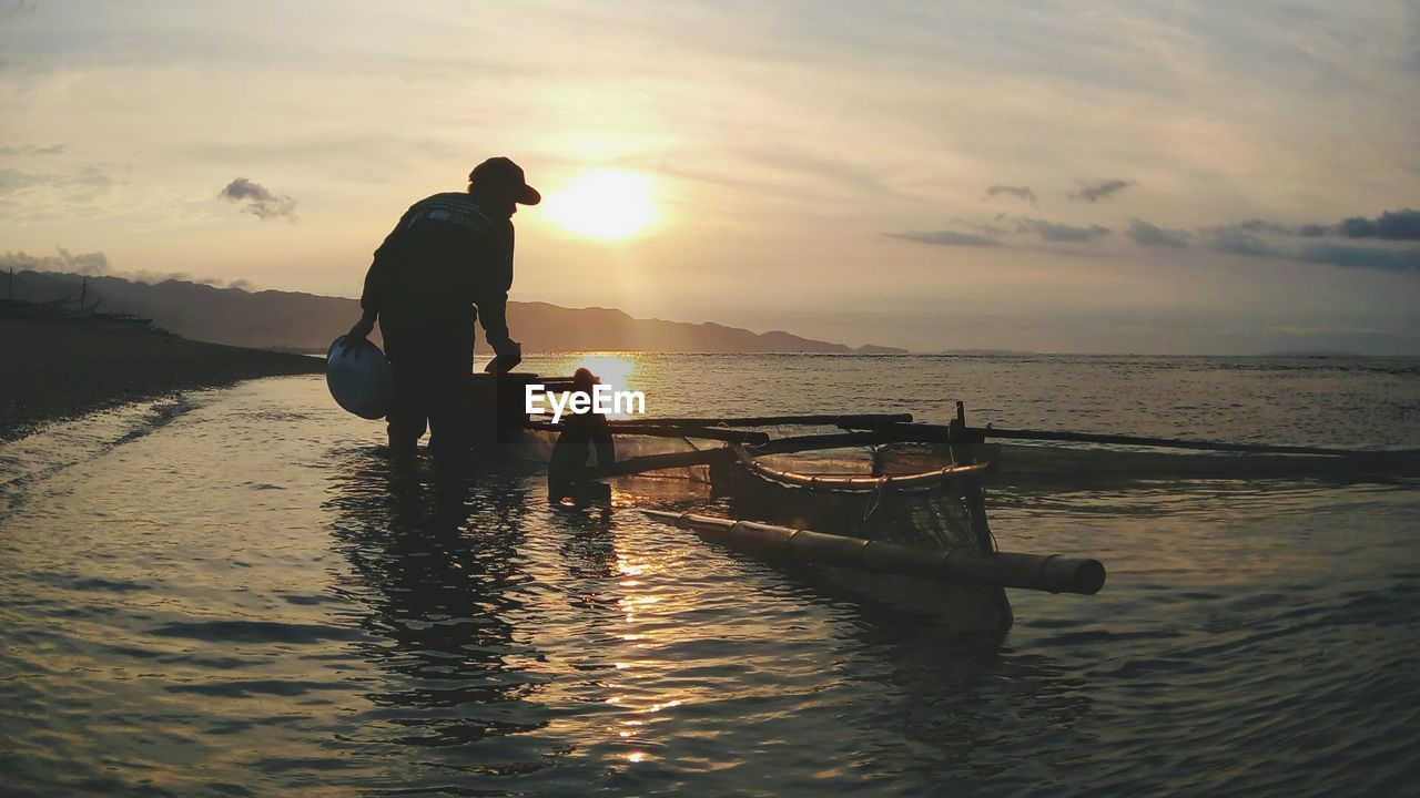 SILHOUETTE MAN STANDING ON BOAT AGAINST SKY DURING SUNSET