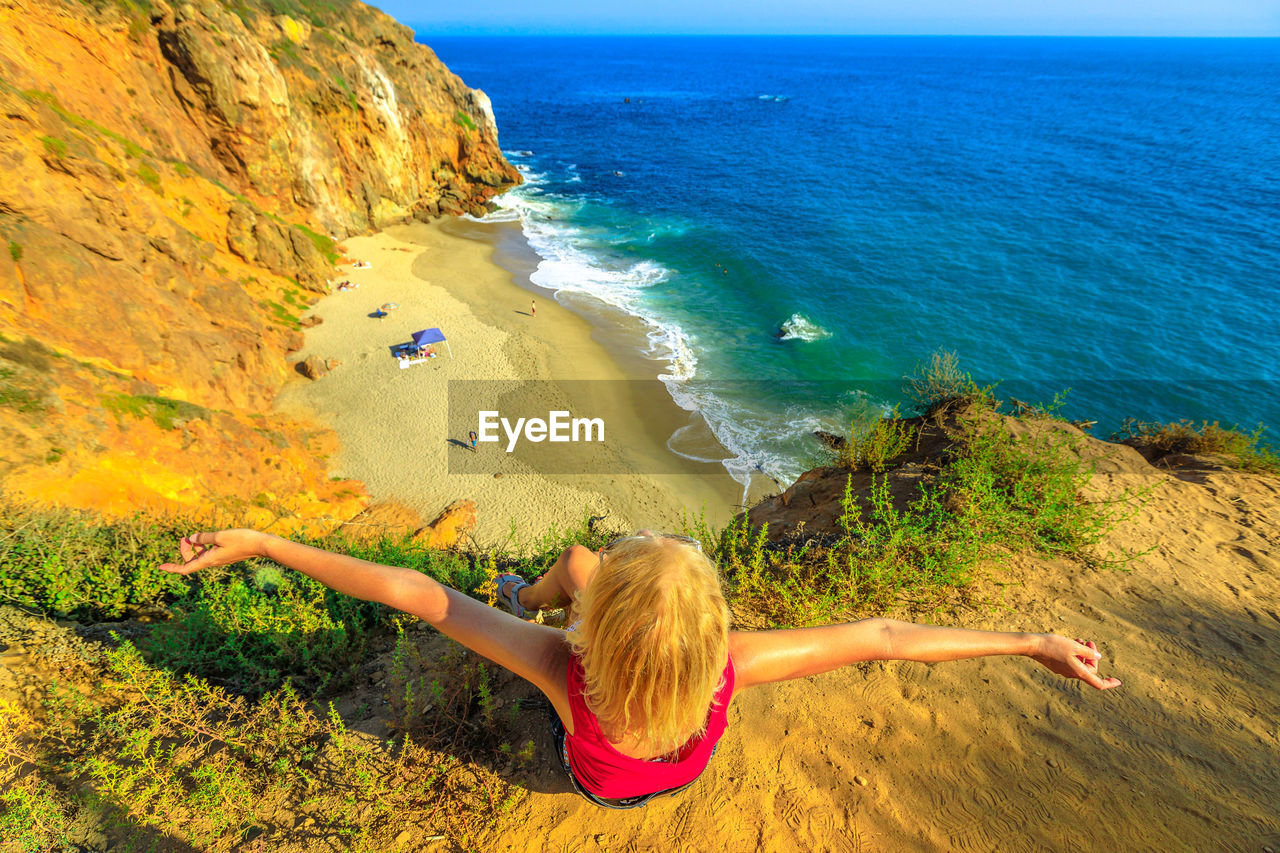 High angle view of mature woman with arms outstretched sitting on cliff by sea