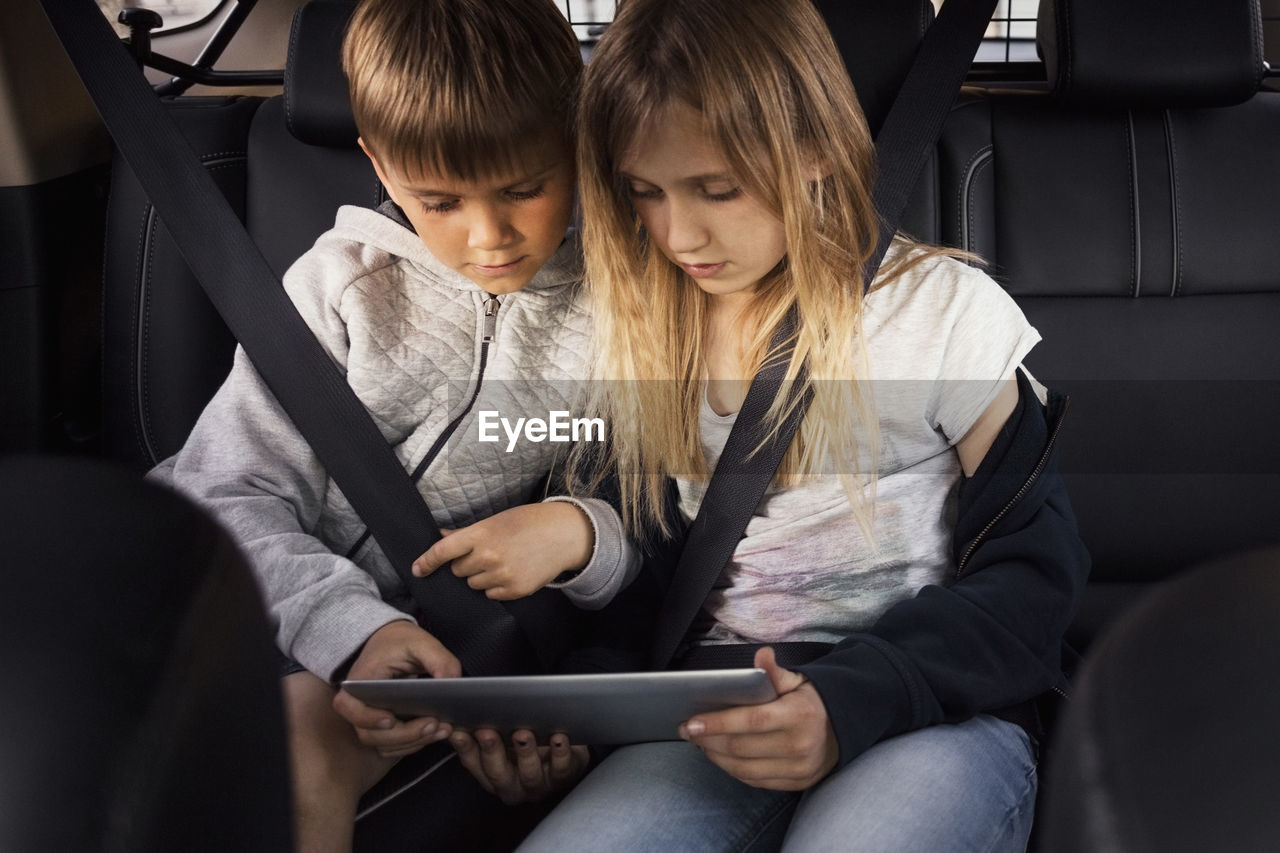 Sibling using digital tablet while sitting in electric car