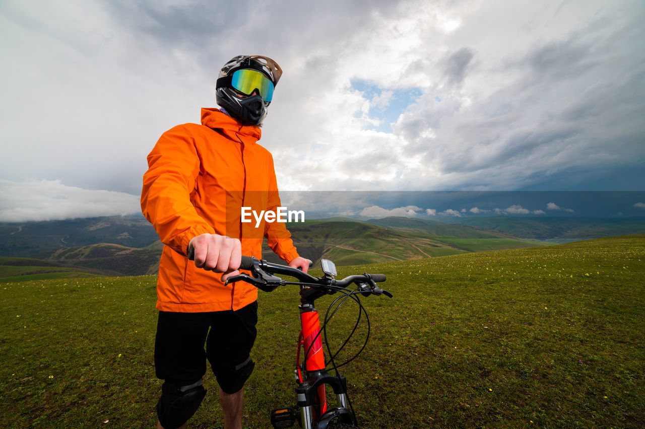 Man in a helmet and protection stands with a bicycle on a green hill among the mountains during a
