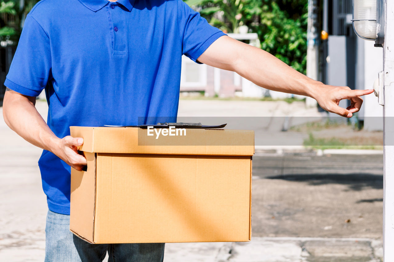 Midsection of delivery person holding cardboard box while ringing doorbell outdoors