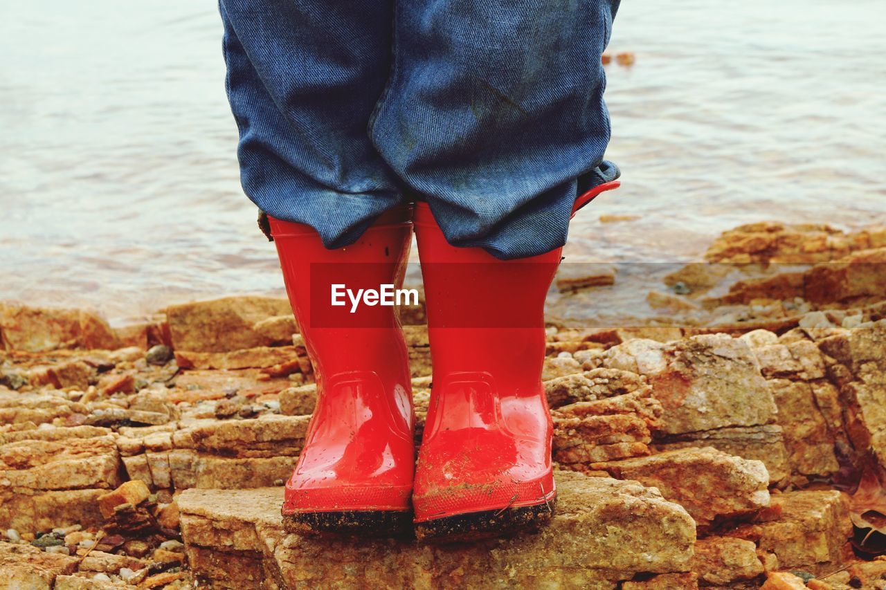 Low section of person wearing red rubber boots while standing on shore