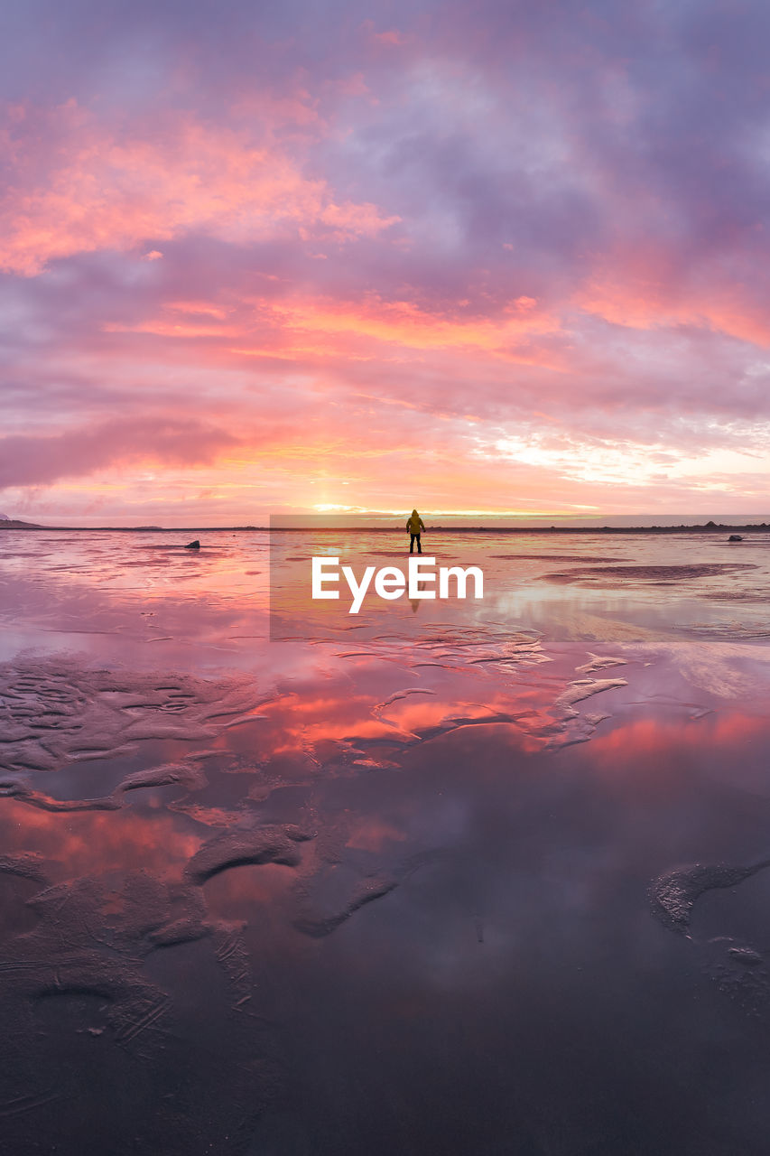 Back view of anonymous tourist standing on wet shore near calm sea during majestic colorful sunset