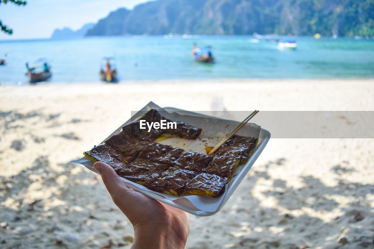 CLOSE-UP OF HAND HOLDING ICE CREAM AGAINST SEA