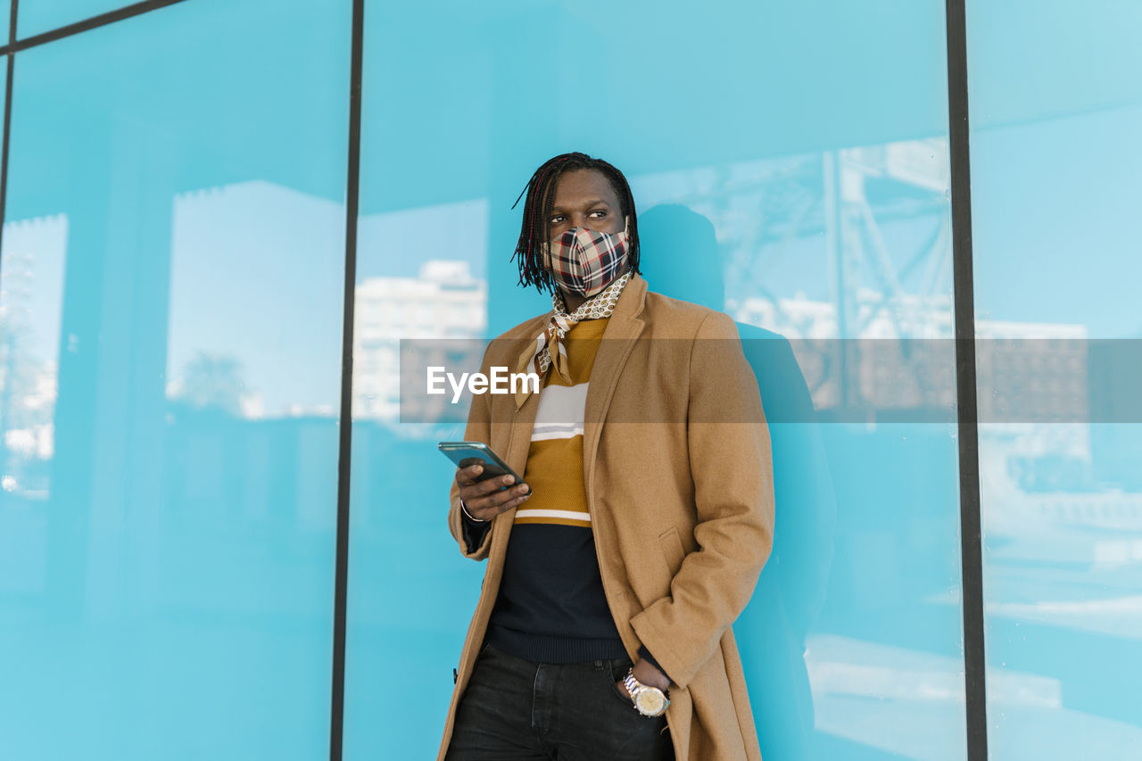 African man in protective face mask with smart phone standing while leaning on blue glass wall