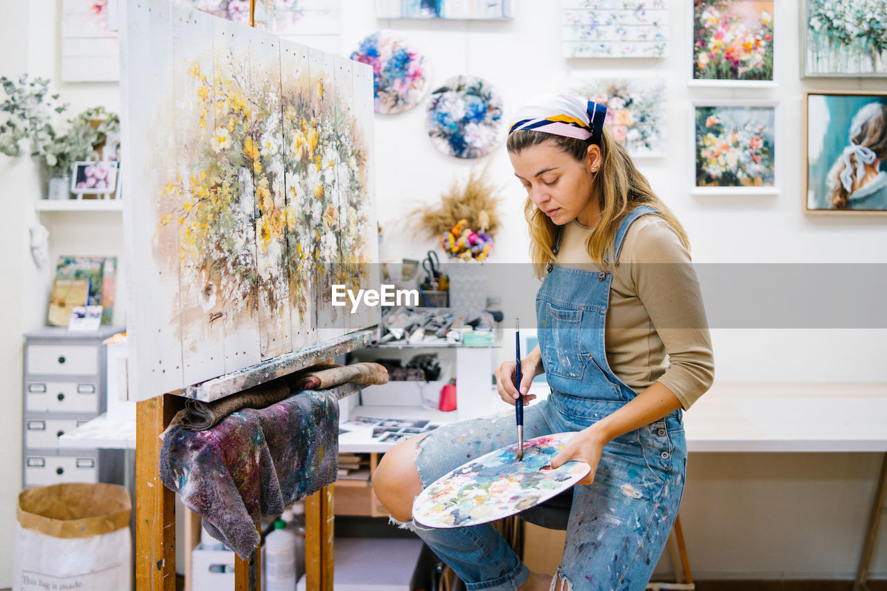 Side view of female artist sitting near easel and drawing colorful picture with paints while working in creative workshop