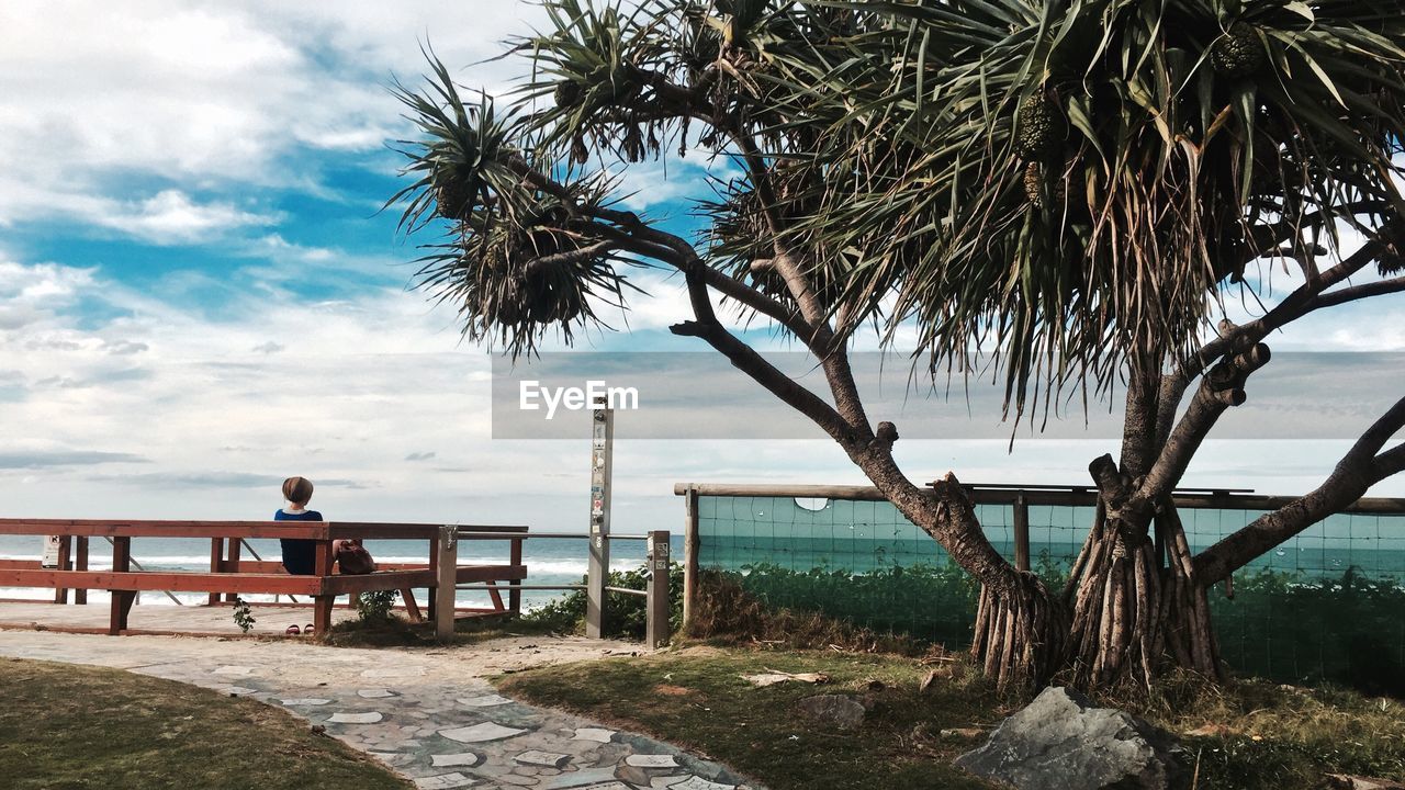 Woman sitting on bench by sea against cloudy sky