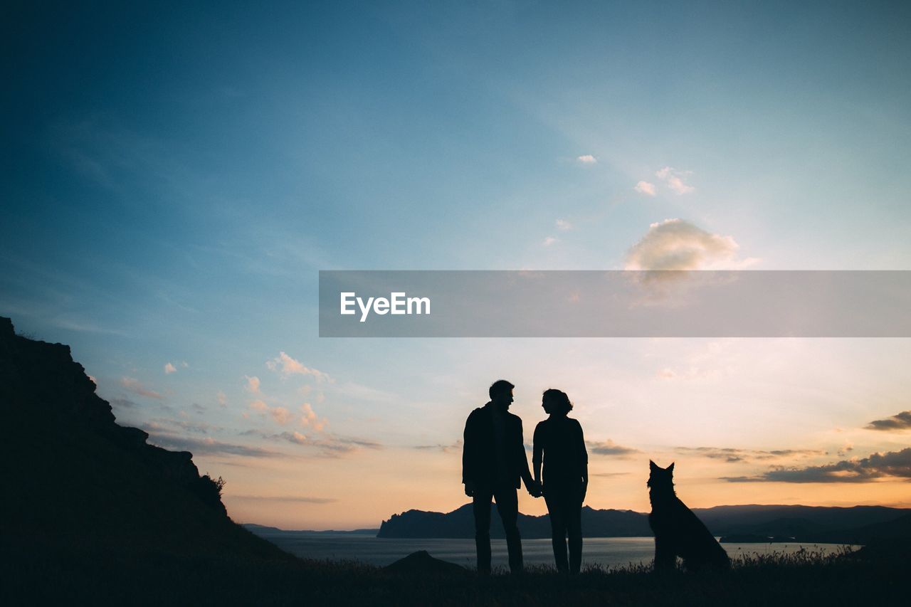 Silhouette couple with dog standing at beach against sky during sunset