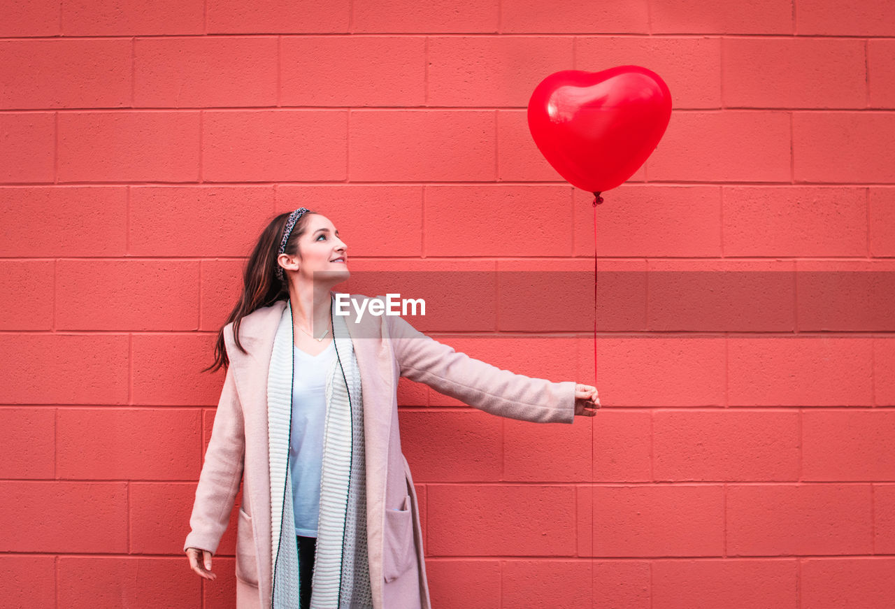 Woman holding heart shape balloon against wall