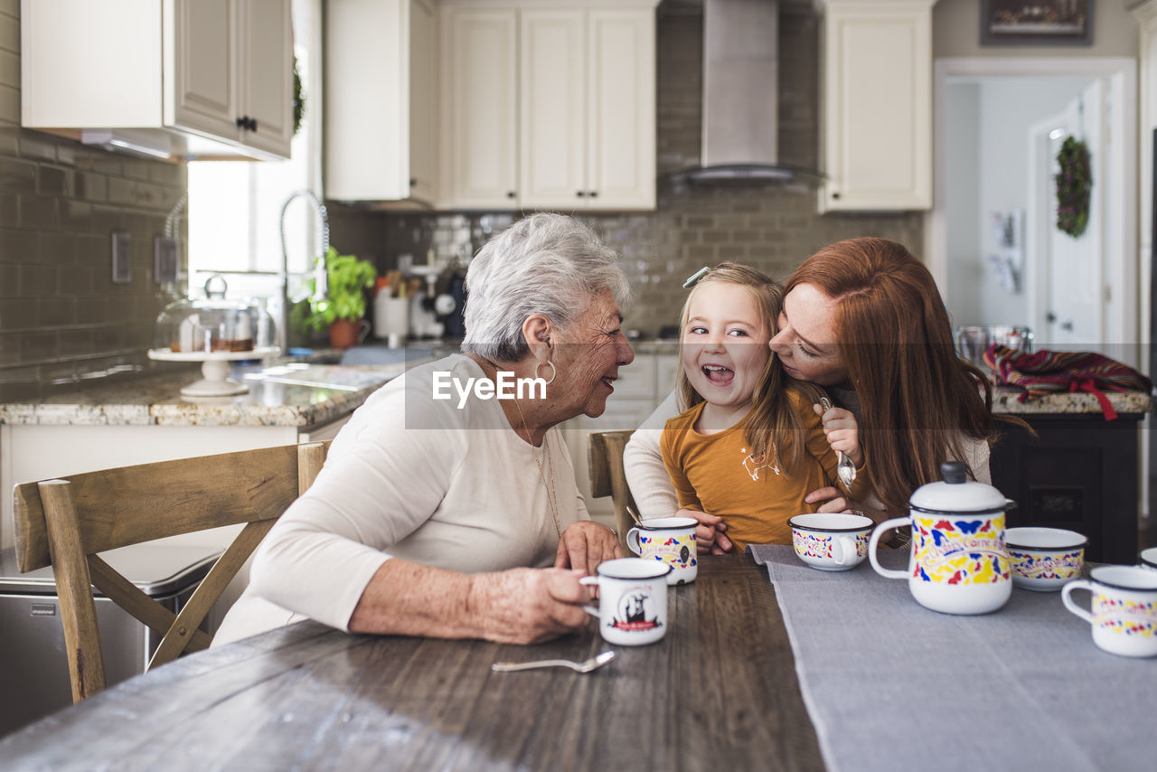 Mother kissing daughter at kitchen table with grandmother looking