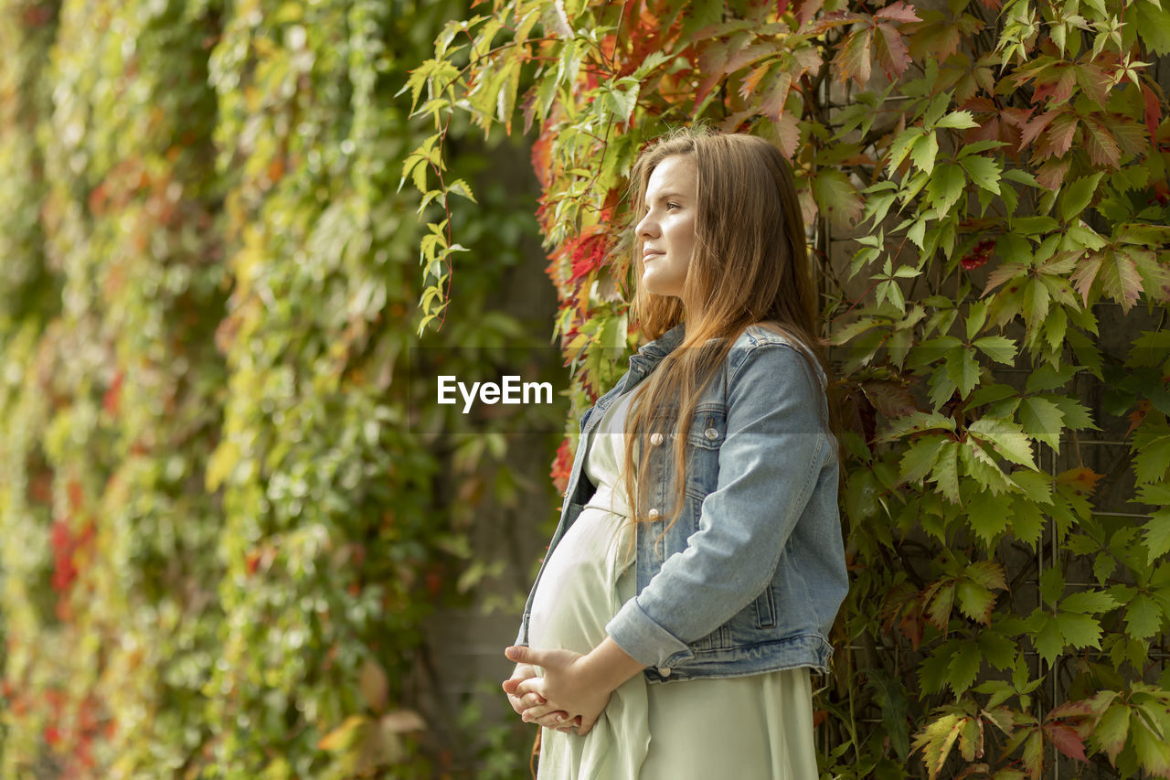 White pretty pregnant woman stand near green overgrown liana wall. long brown haired young healthy