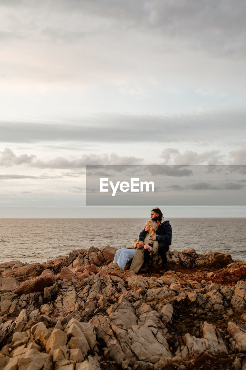 Bearded man embracing blond girlfriend in outerwear and looking away while sitting on stony coast of rippling sea on cloudy day in aviles, spain