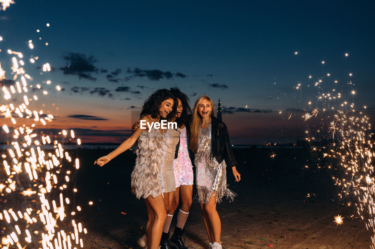 Cheerful young female friends with firework at beach against sky during sunset