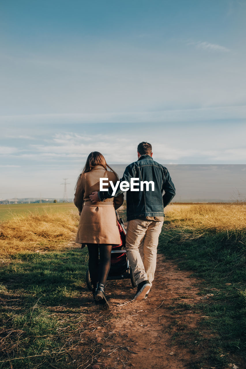 Back view of tender couple hugging and walking with baby stroller along road in countryside on sunny day