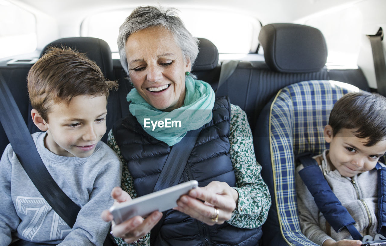 Happy senior woman using smart phone with grandsons in car