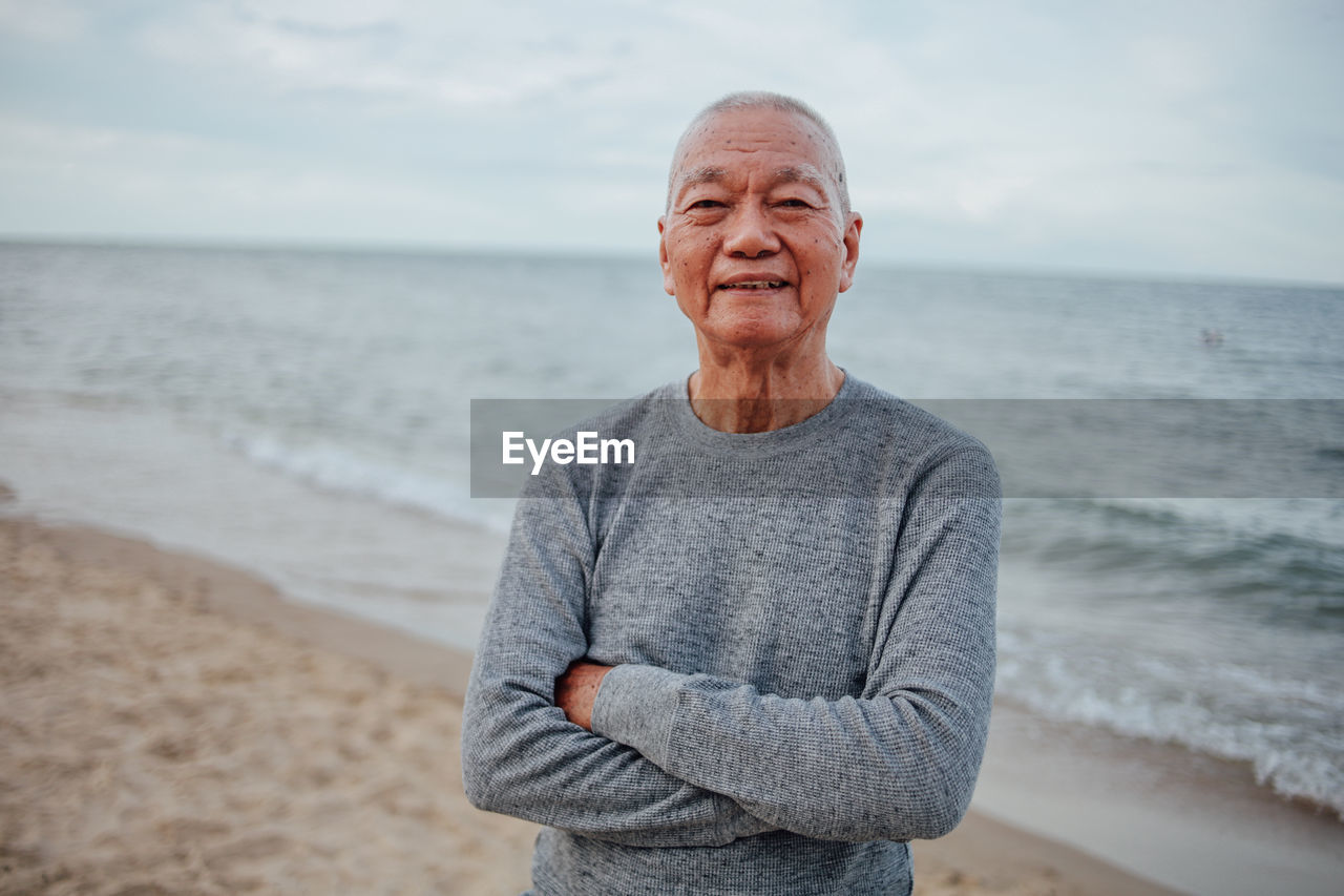 Portrait of senior man with arms crossed standing at beach against sky during sunset