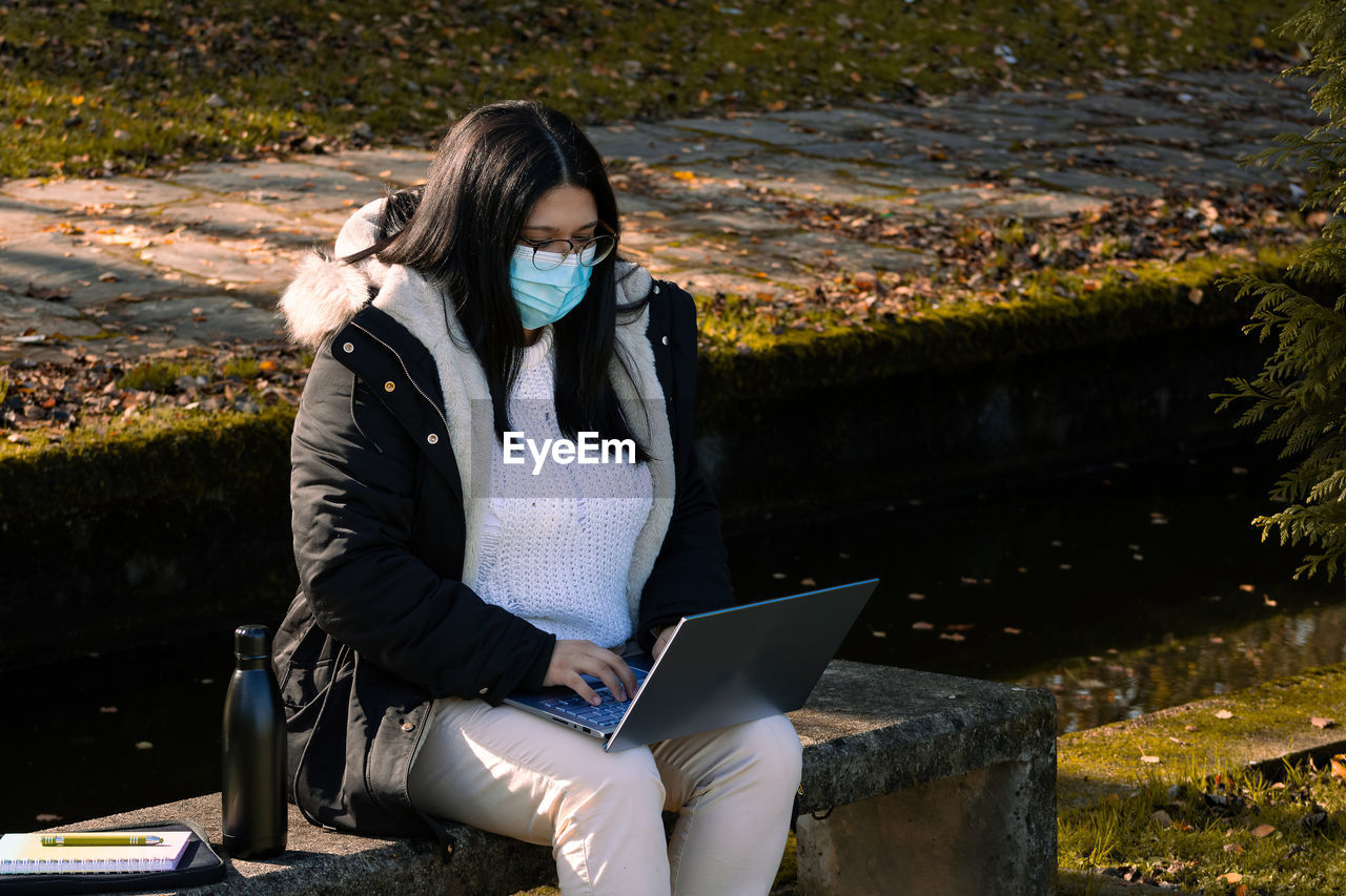 Woman with surgical mask working in the park with laptop. teleworking outdoors due to coronavirus.