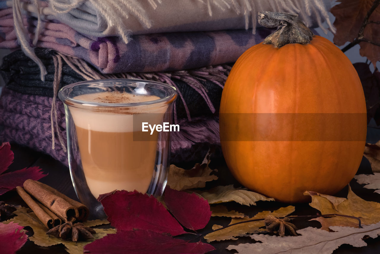 Heat-resistant glass cup with pumpkin latte and  pumpkin stand on table with stack of scarves