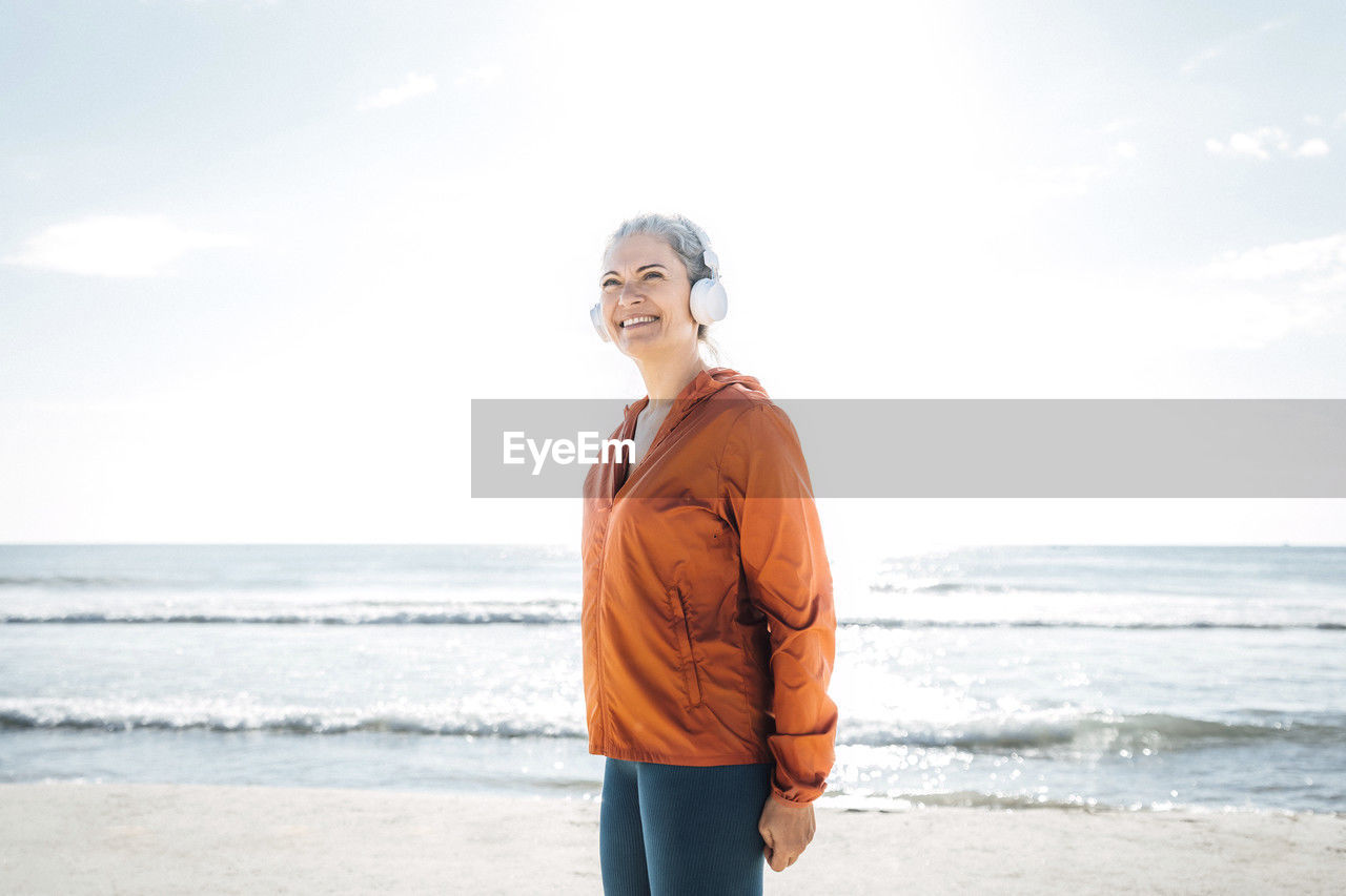 Smiling mature woman wearing headphones listening to music at beach