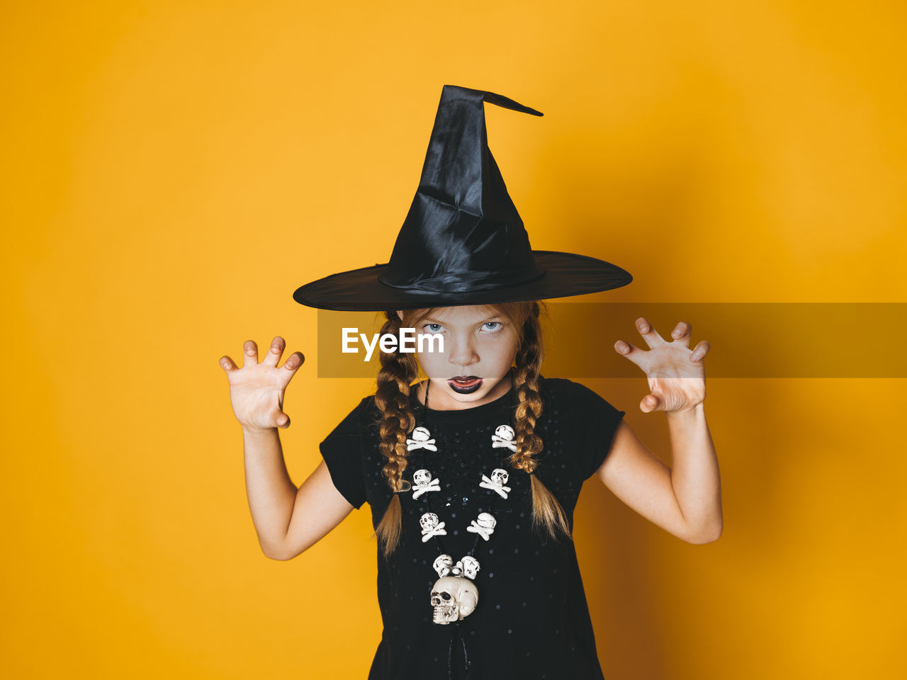 Portrait of girl wearing witch hat gesturing against yellow background during halloween