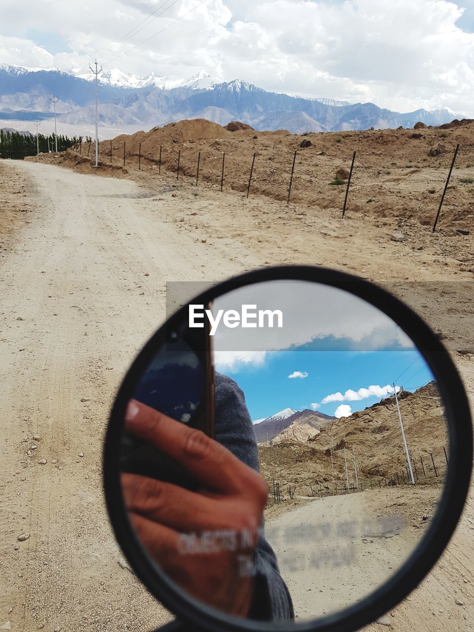 Reflection of man photographing with mobile phone in side-view mirror on dirt road