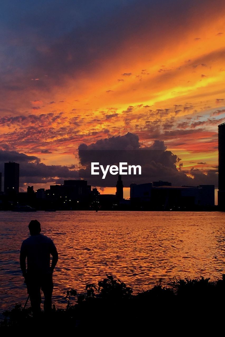 Silhouette man standing by sea against cloudy orange sky in city during sunset