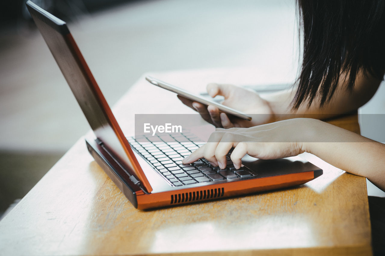 Cropped hands of woman using smart phone and laptop on wooden table