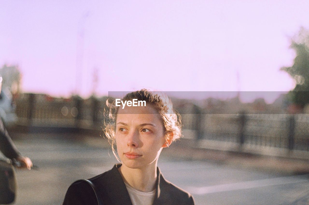 Portrait of young woman looking away in city against sky