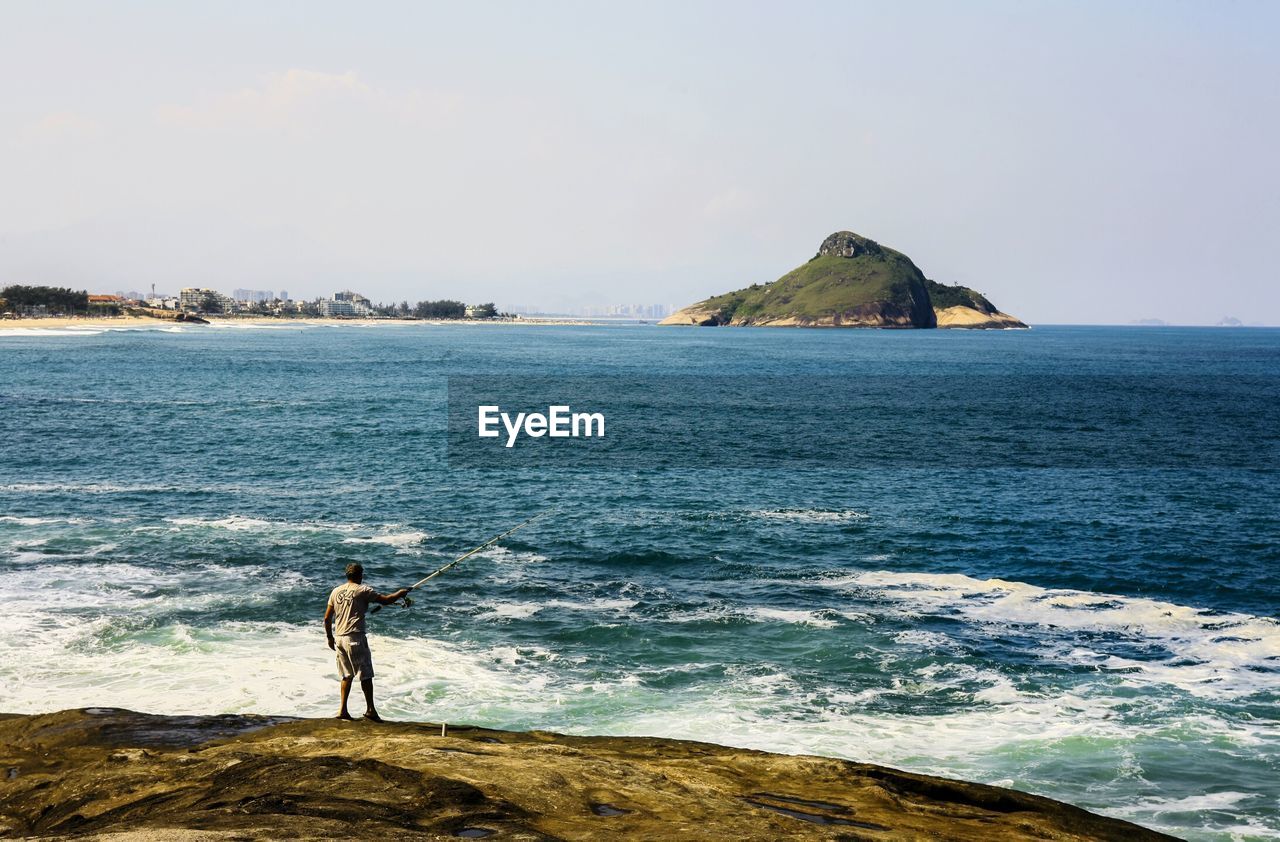 MAN STANDING ON ROCK AGAINST SEA