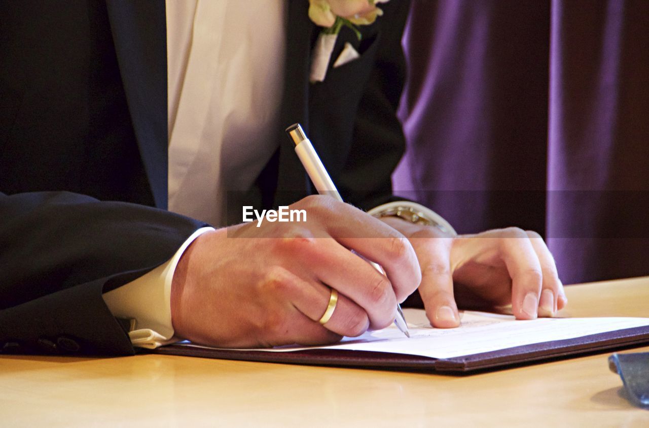 Midsection of bridegroom signing document on table during wedding