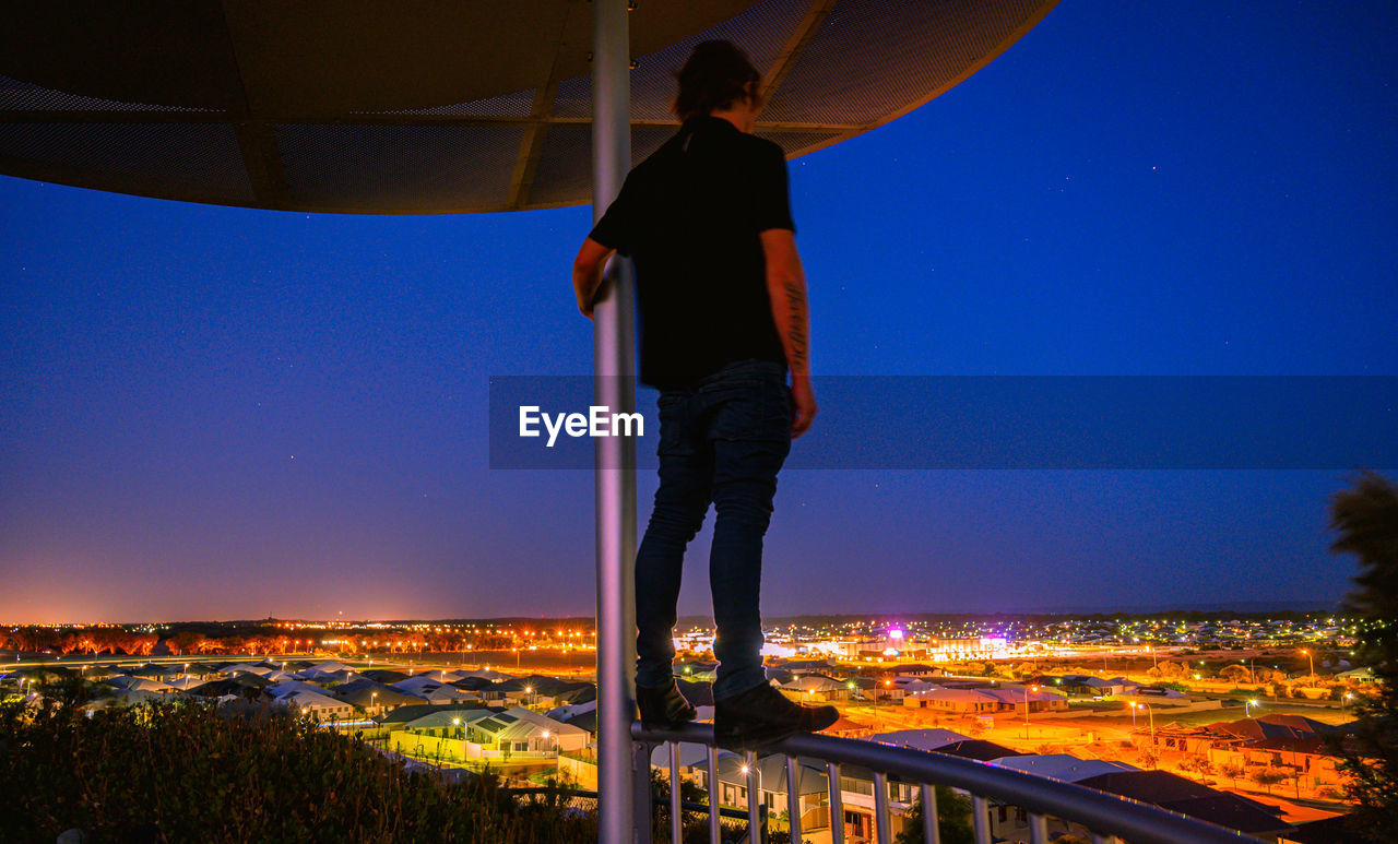 Man standing by illuminated city against clear blue sky at night