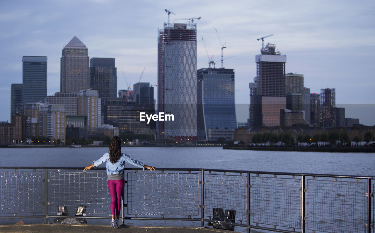 Rear view of woman standing by river with buildings in background